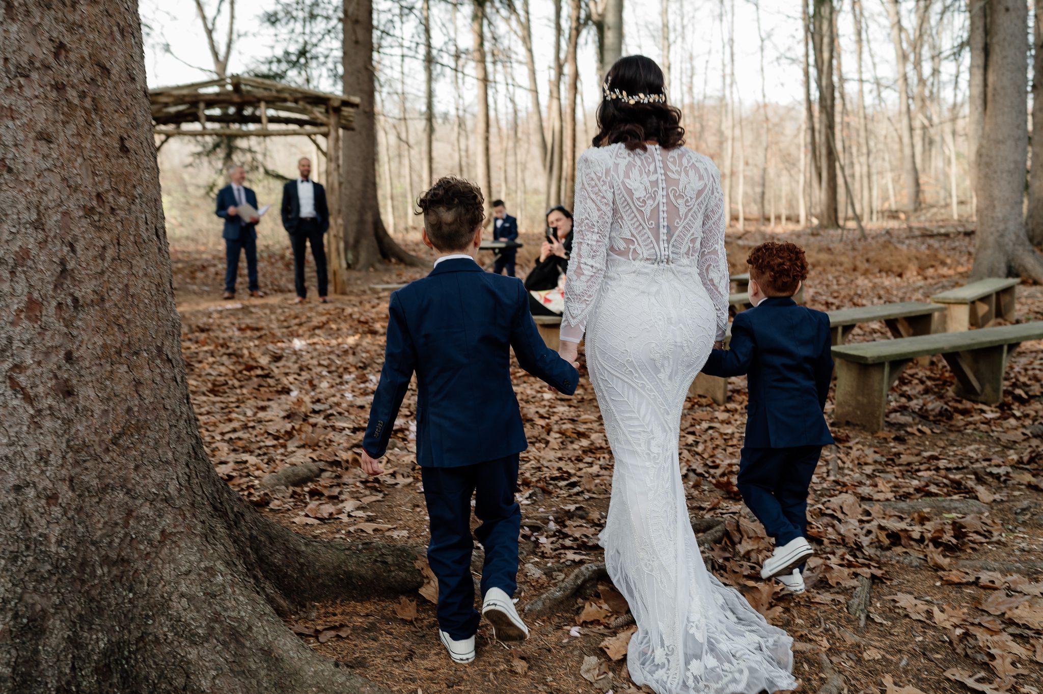 bride in white gown walks down aisle of leaves through woods with her 2 young sons