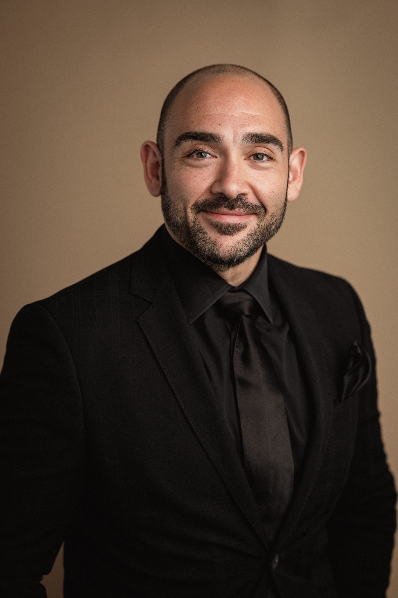 man in black suit poses in front of tan backdrop for headshot