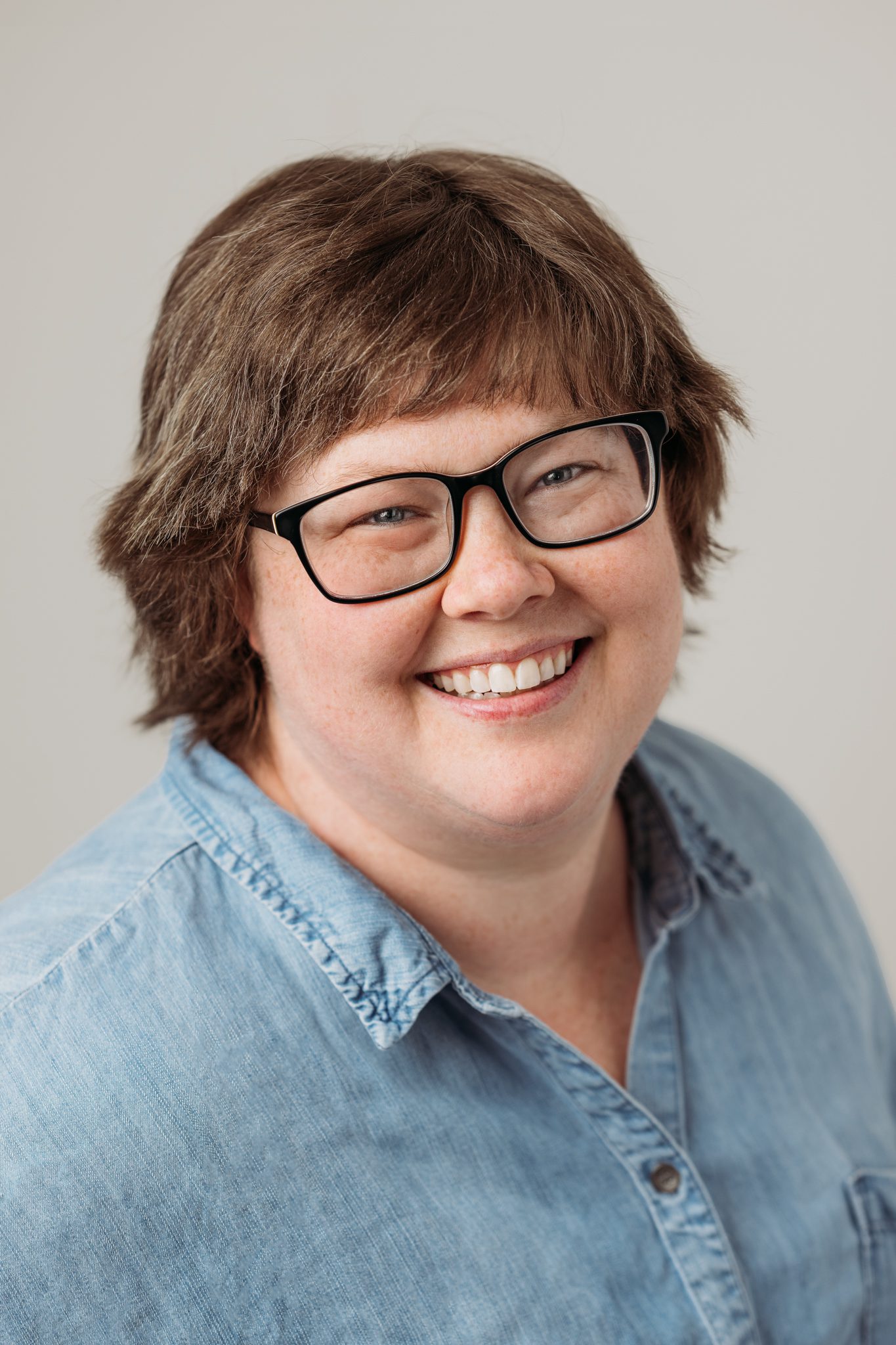 woman in blue shirt wearing glasses poses for headshot in ront of a white backdrop