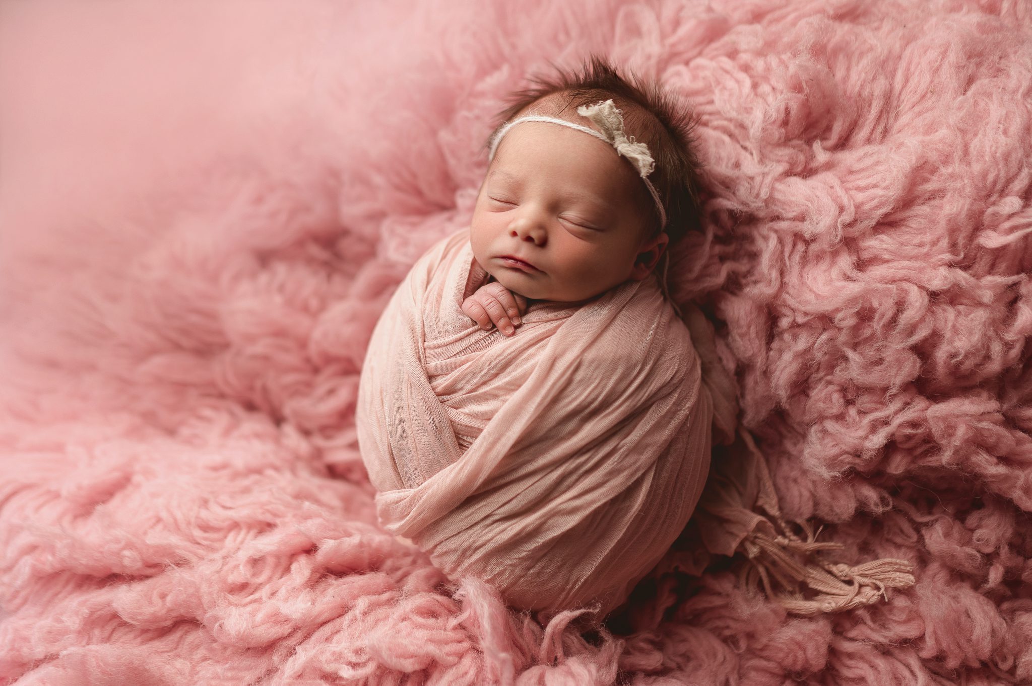 A tiny newborn baby girl wrapped in a soft pink swaddle during a mini newborn session in a Scotch Plains photo studio.