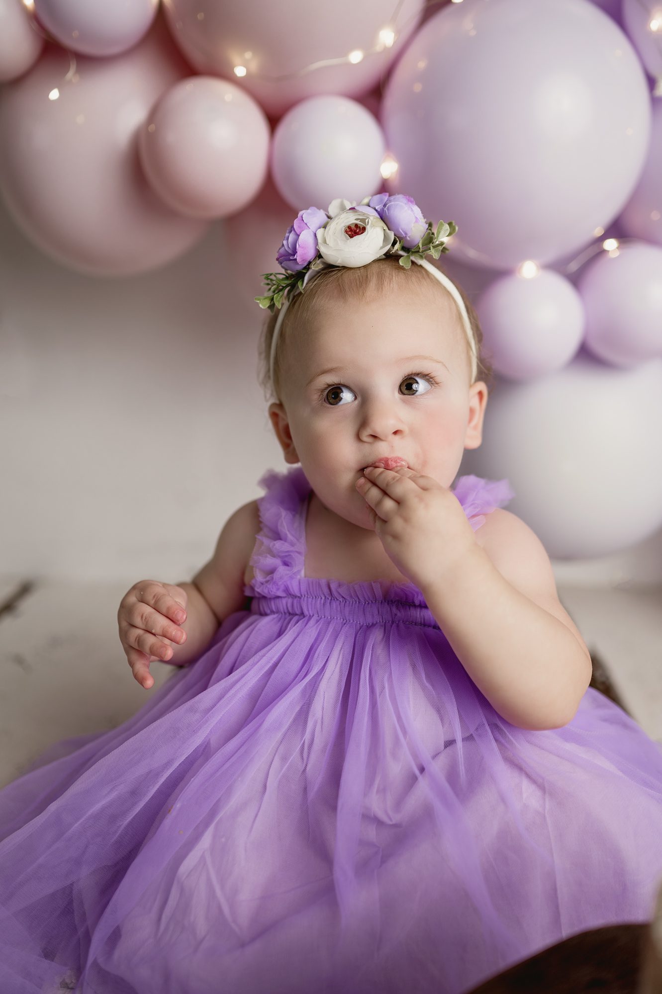 1 year old girl poses for her first birthday portraits with purple balloons in background