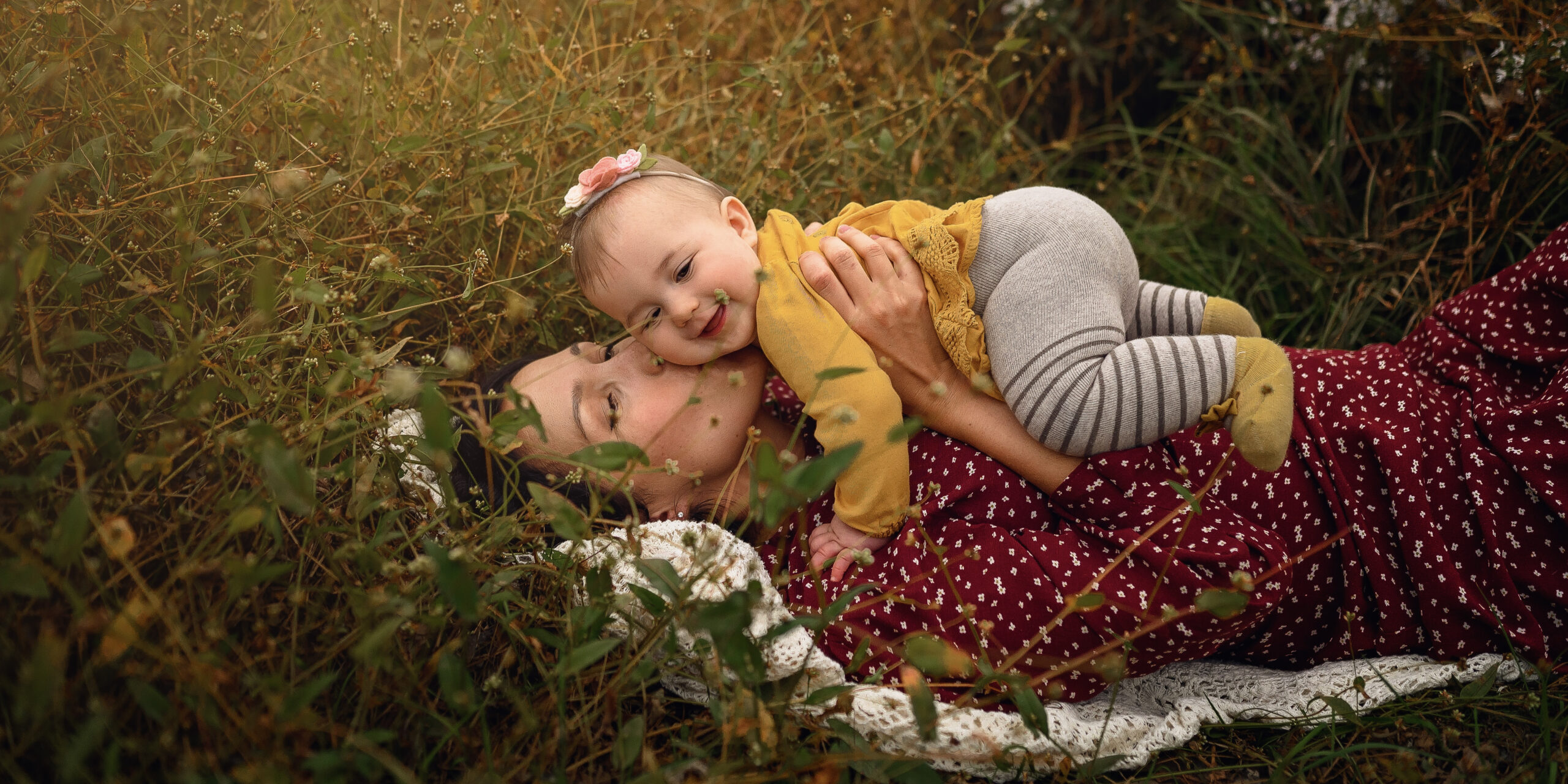 mother lies in a field wearing a red dress and holding her little baby girl in an embrace