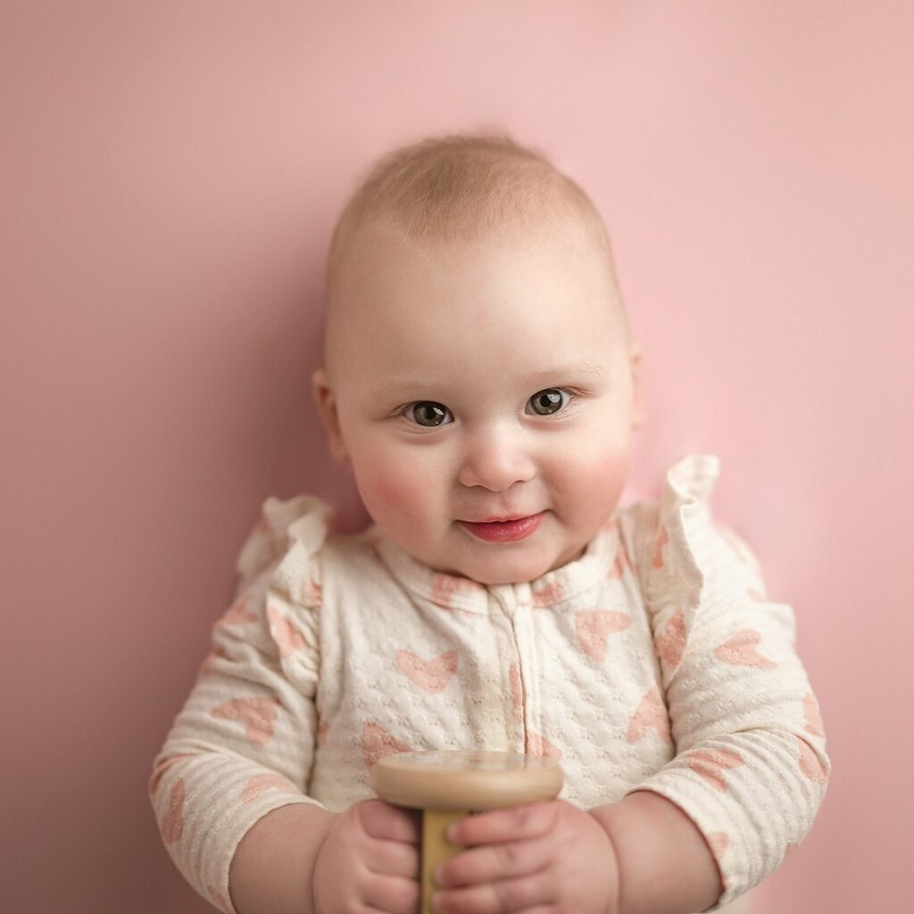adorable six month old with ruffle sleeve cream onesie decorated with pink heards laying on pink backdrop