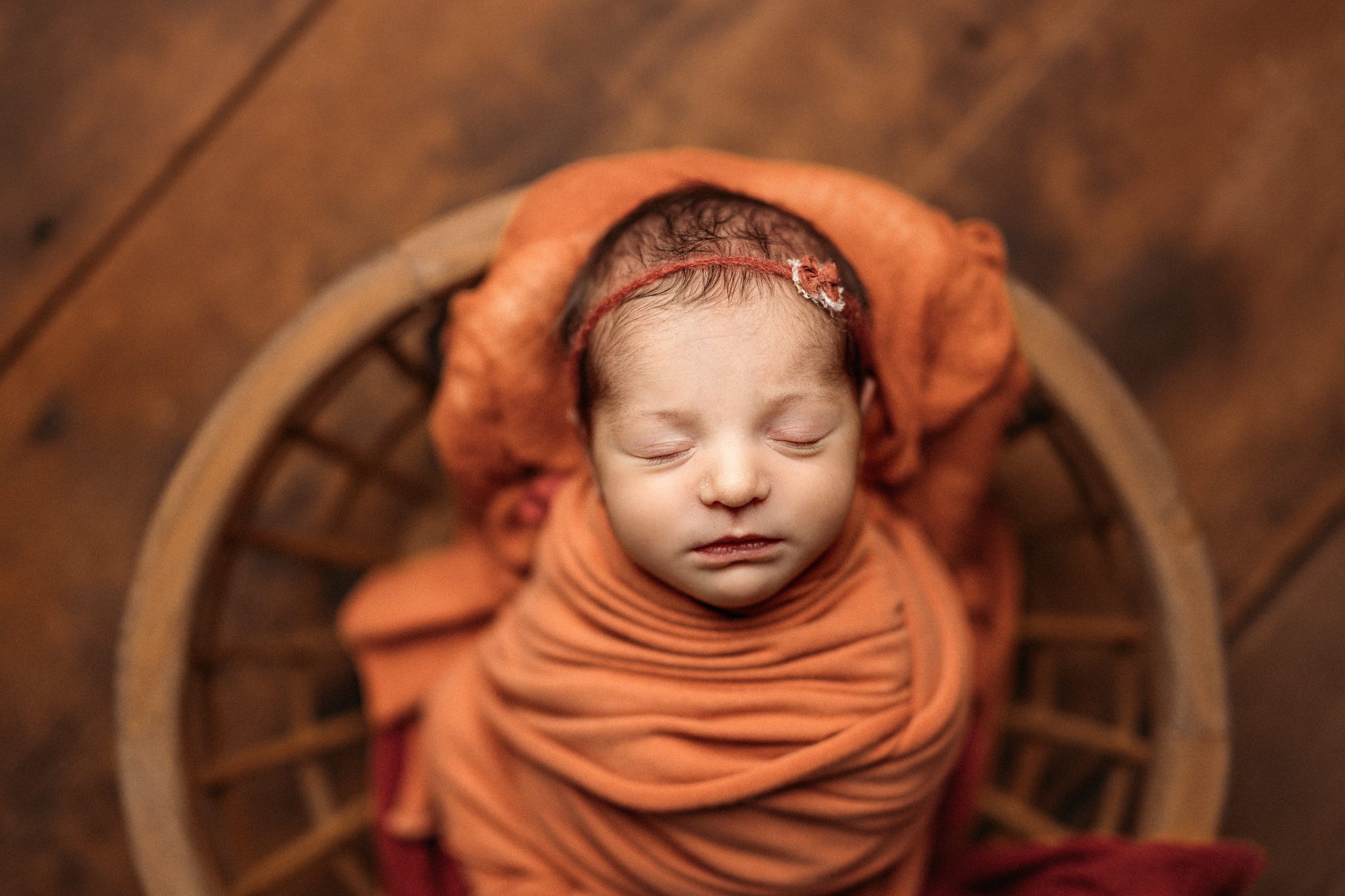 newborn baby girl in rust colored fabric and little bow sleeping in a wooden bowl