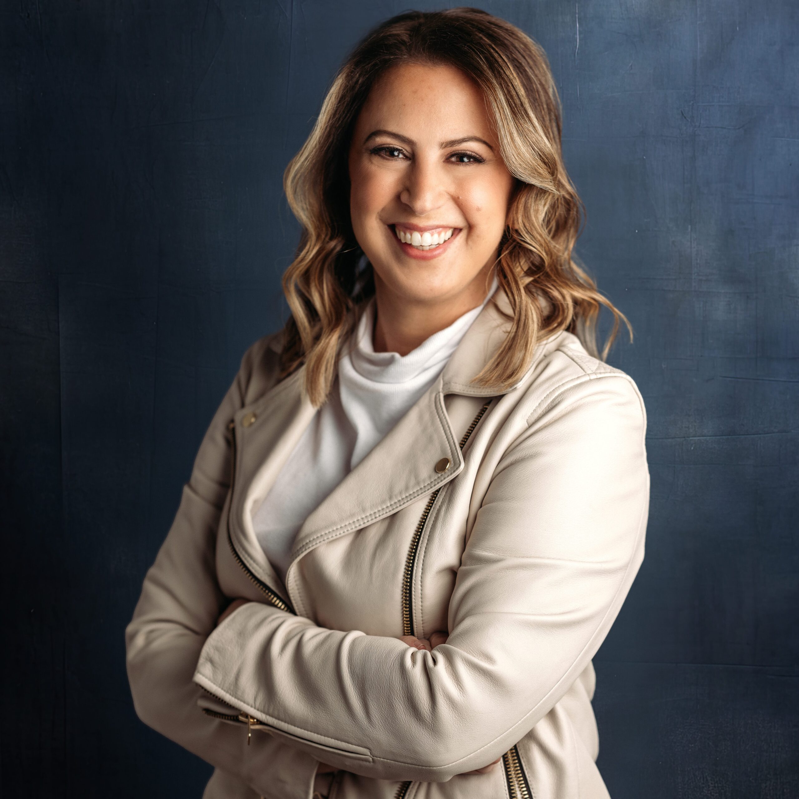 woman with brown highlighted beach wave hair wearing a cream leather jacket poses for a headshot photo on navy background