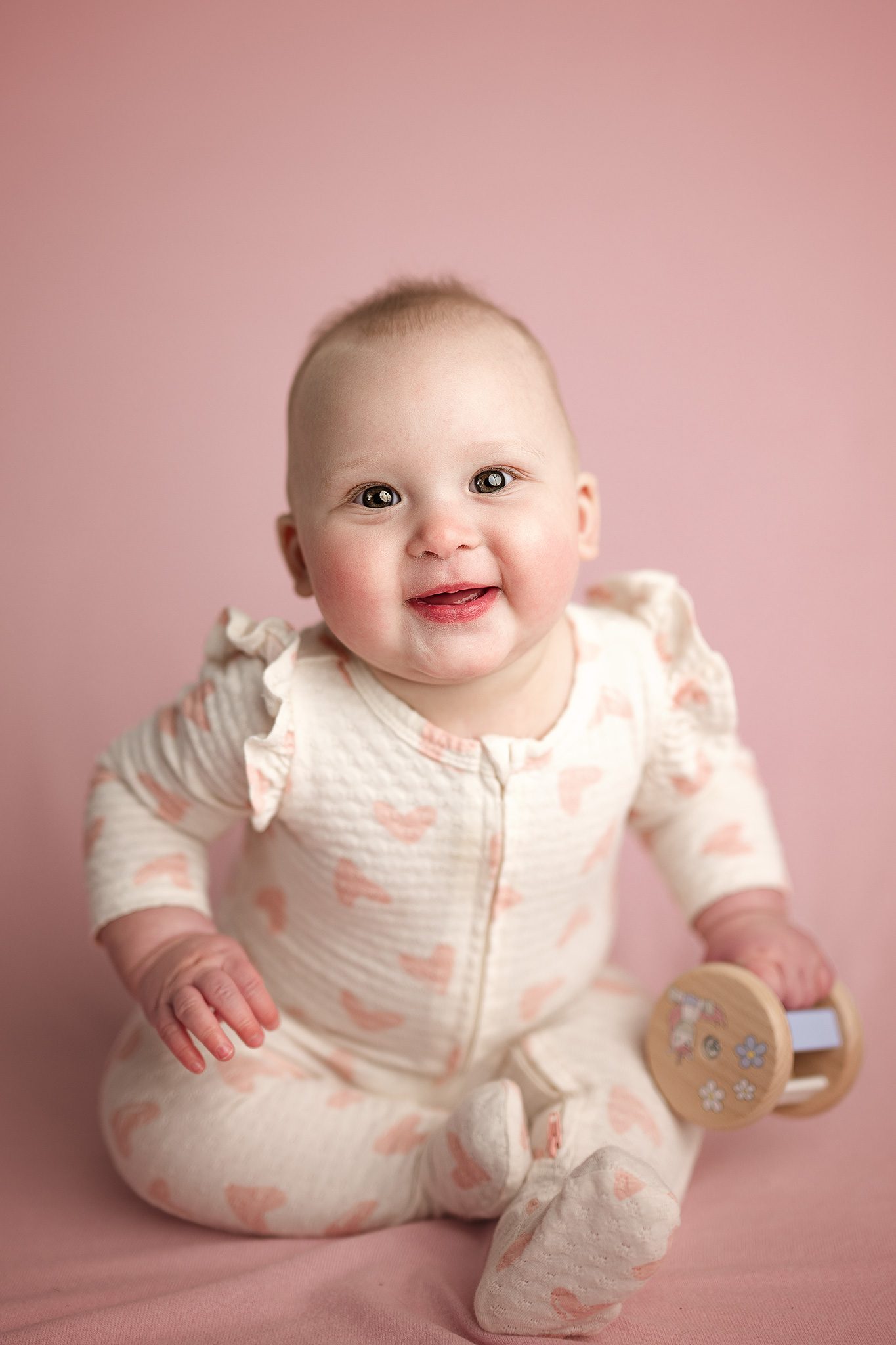 6 month old baby sits up and smiles on a pink backdrop in her cream ruffle sleeved pajamas decorated in little pink hearts