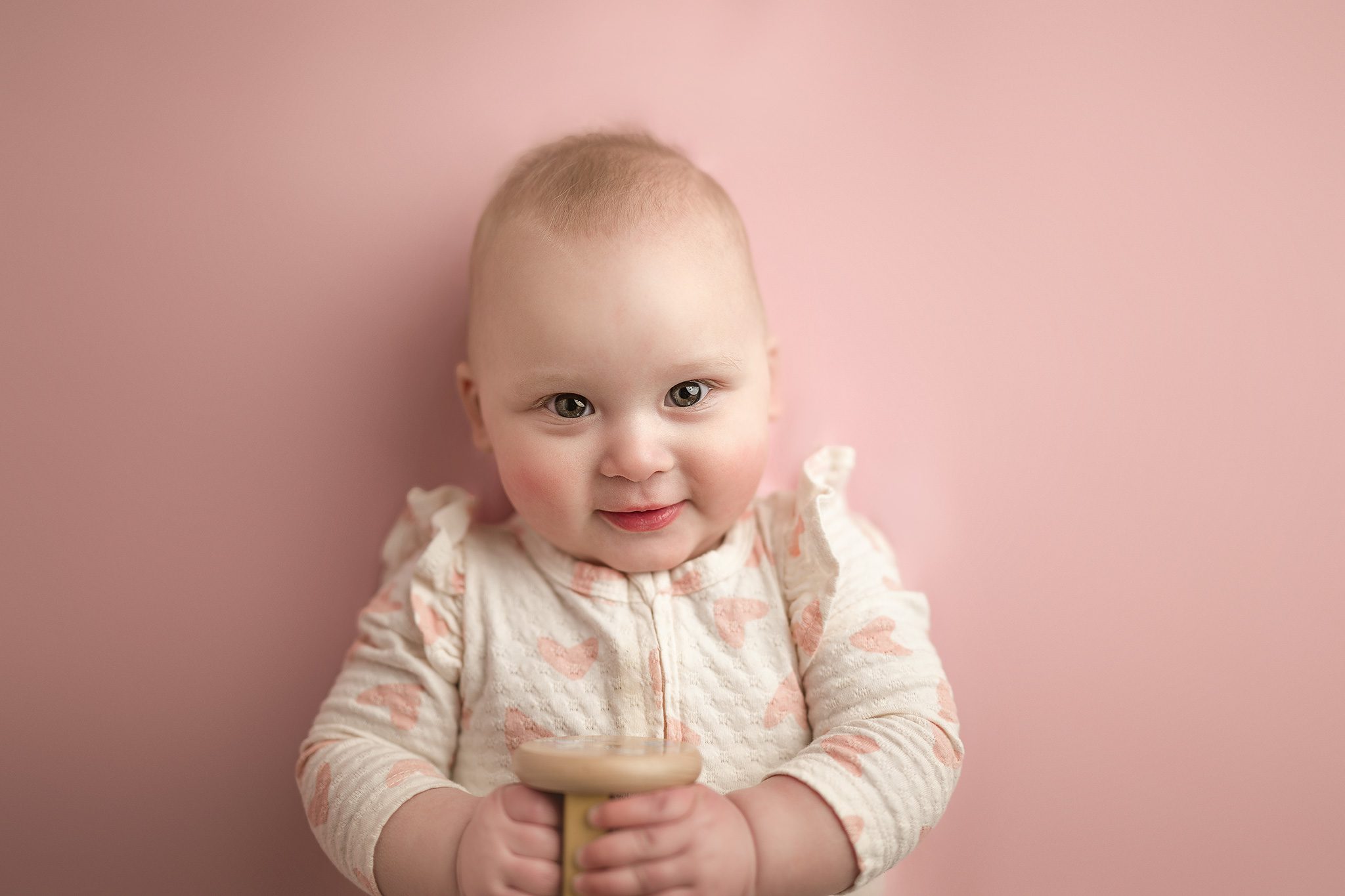 6 month old baby sits up and smiles on a pink backdrop in her cream ruffle sleeved pajamas decorated in little pink hearts