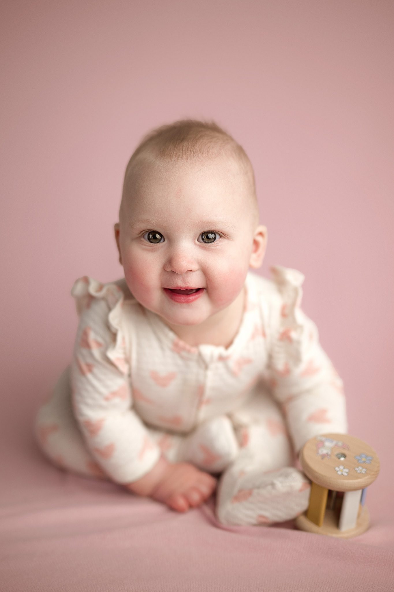 6 month old baby sits up and smiles on a pink backdrop in her cream ruffle sleeved pajamas decorated in little pink hearts