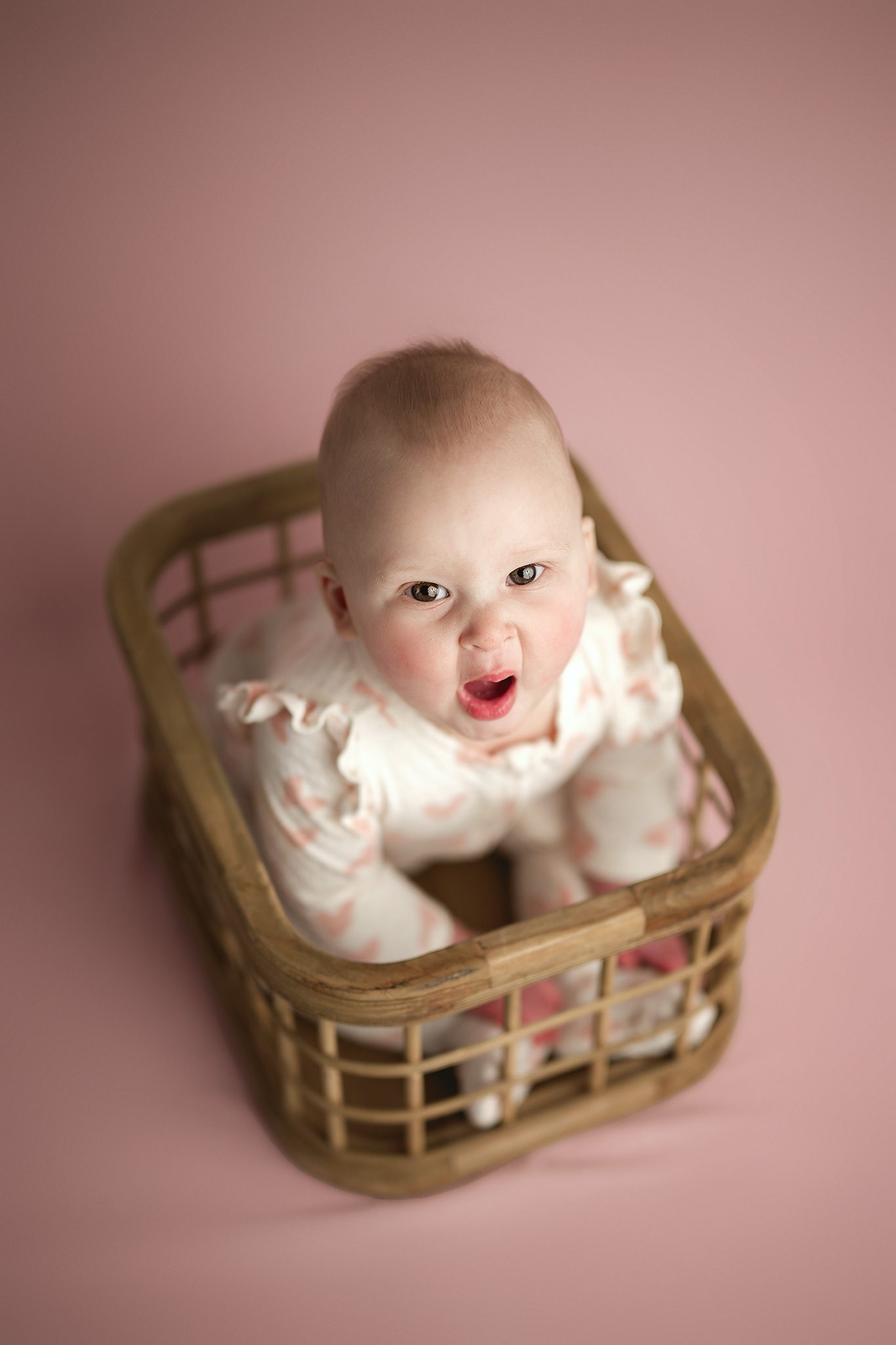 6 month old baby sits up and smiles on a pink backdrop in her cream ruffle sleeved pajamas decorated in little pink hearts