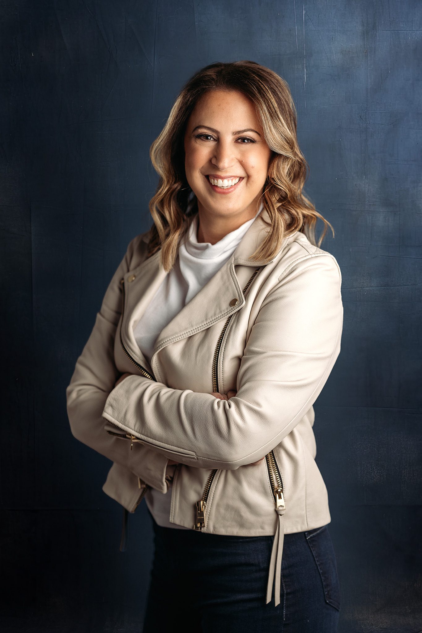 woman with brown highlighted beach wave hair wearing a cream leather jacket poses for a headshot photo on navy background
