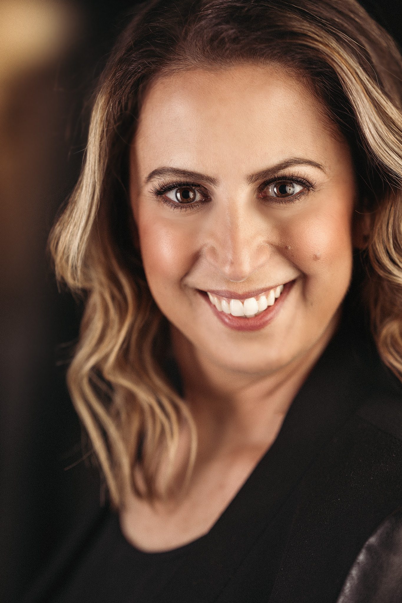 woman with brown highlighted beach wave hair wearing a black leather jacket poses for a headshot photo