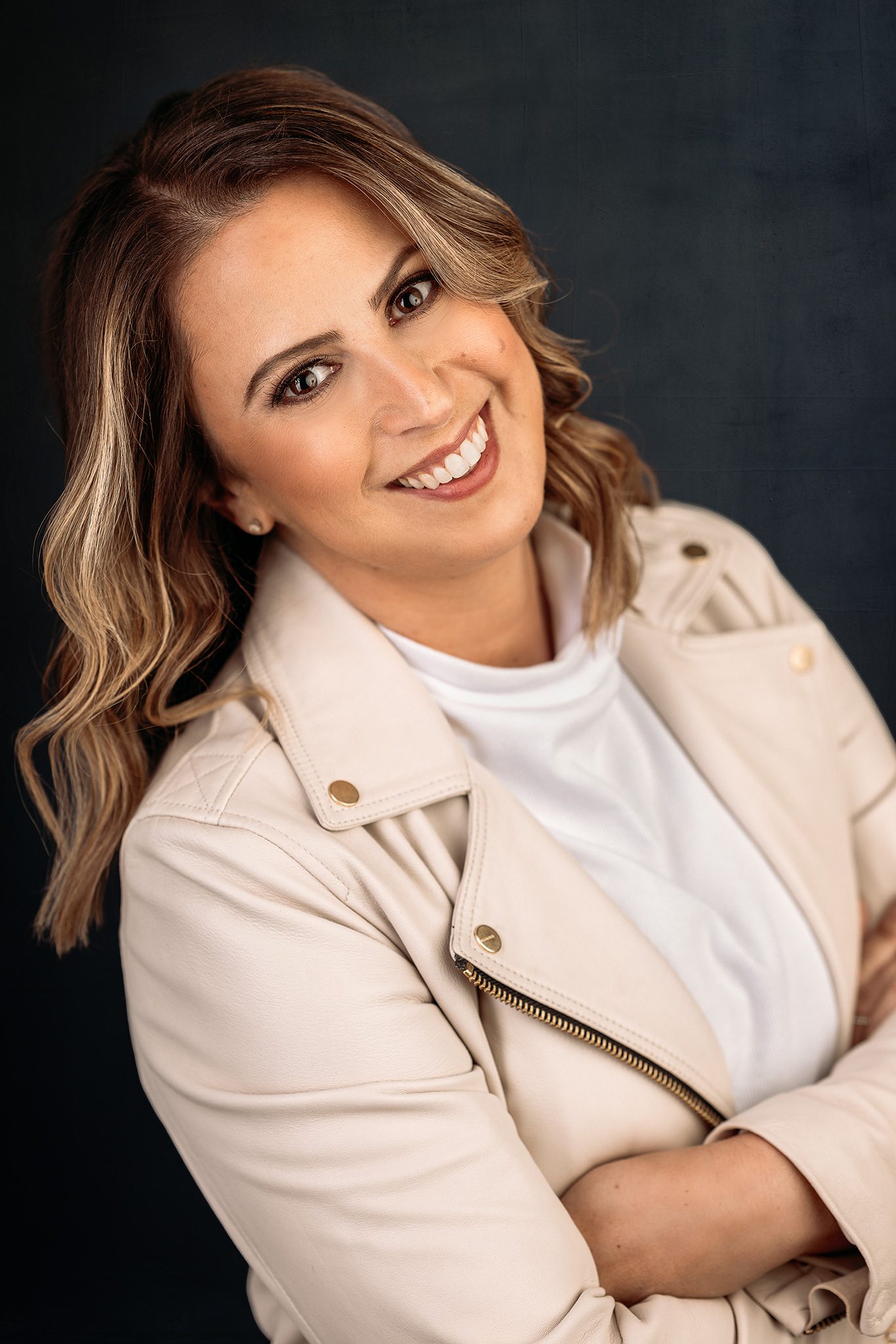 woman with brown highlighted beach wave hair wearing a cream leather jacket poses for a headshot photo on navy background