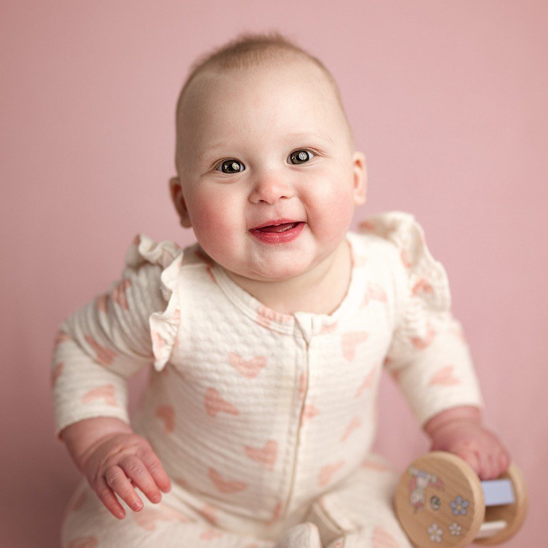 6 month old baby sits up and smiles on a pink backdrop in her cream ruffle sleeved pajamas decorated in little pink hearts