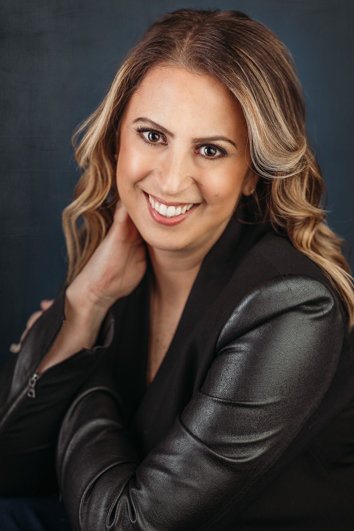 woman with brown highlighted beach wave hair wearing a black leather jacket poses for a headshot photo on navy background
