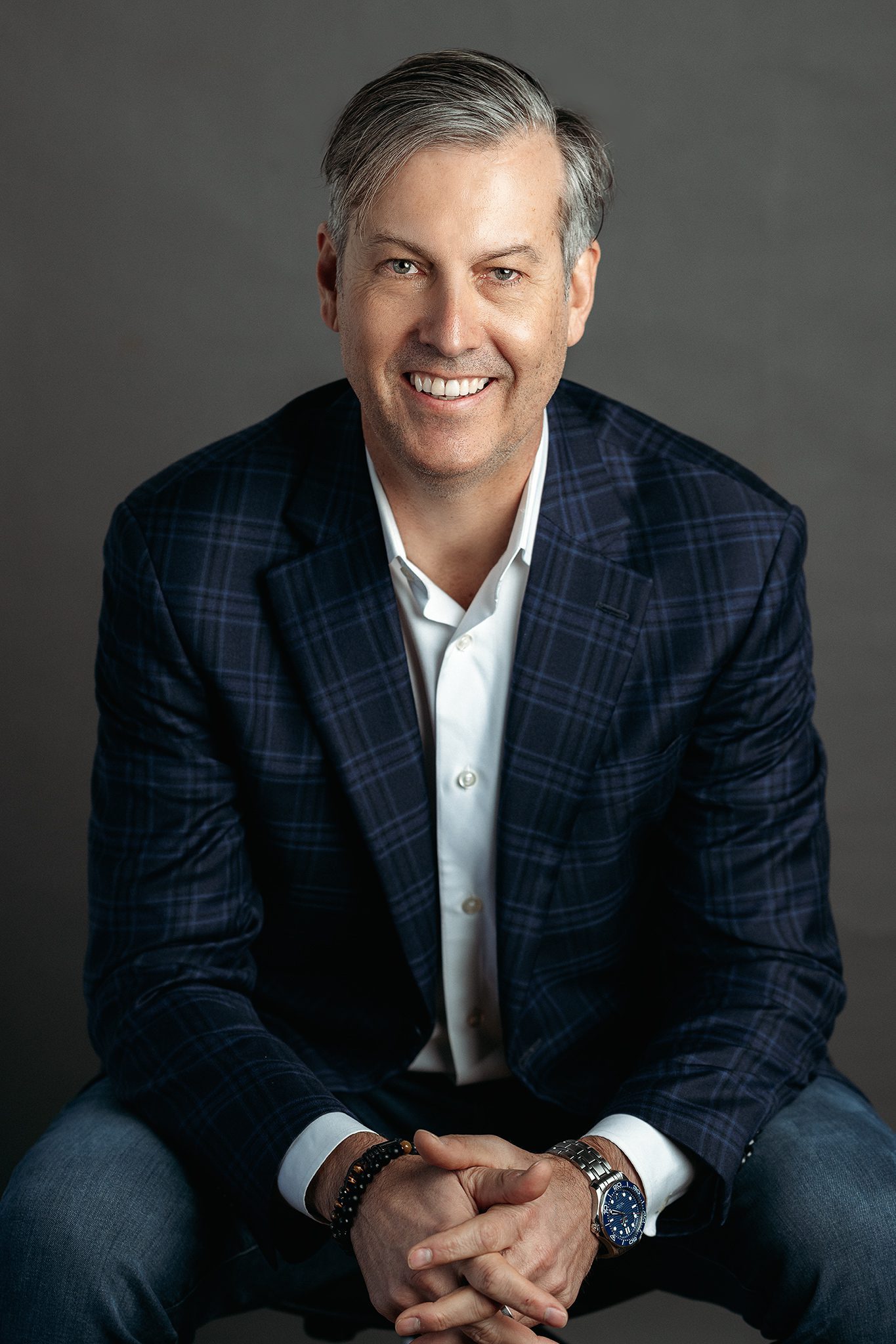 man wearing a blue blazer in jeans poses for headshot on grey backdrop