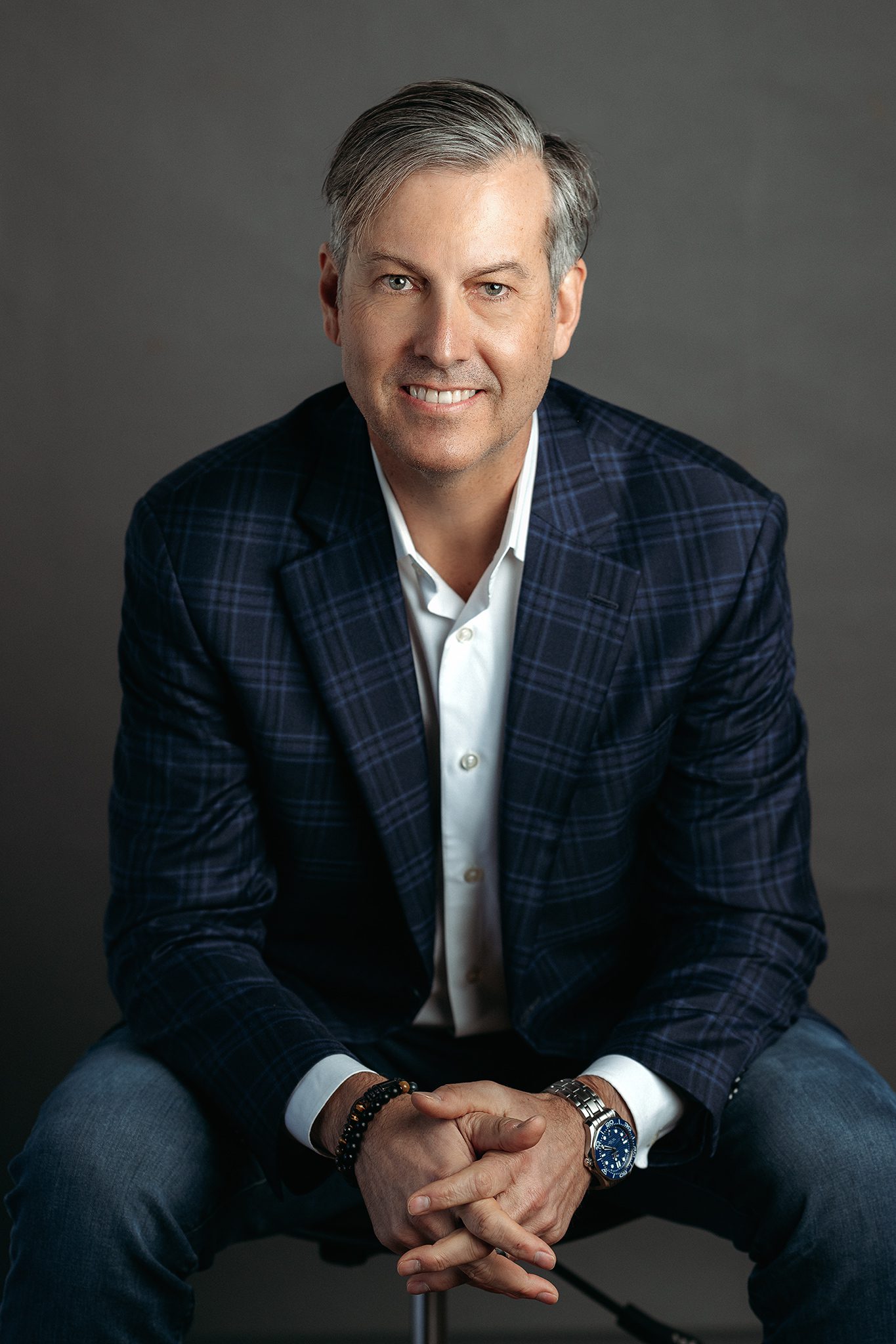 man wearing a blue blazer in jeans poses for headshot on grey backdrop