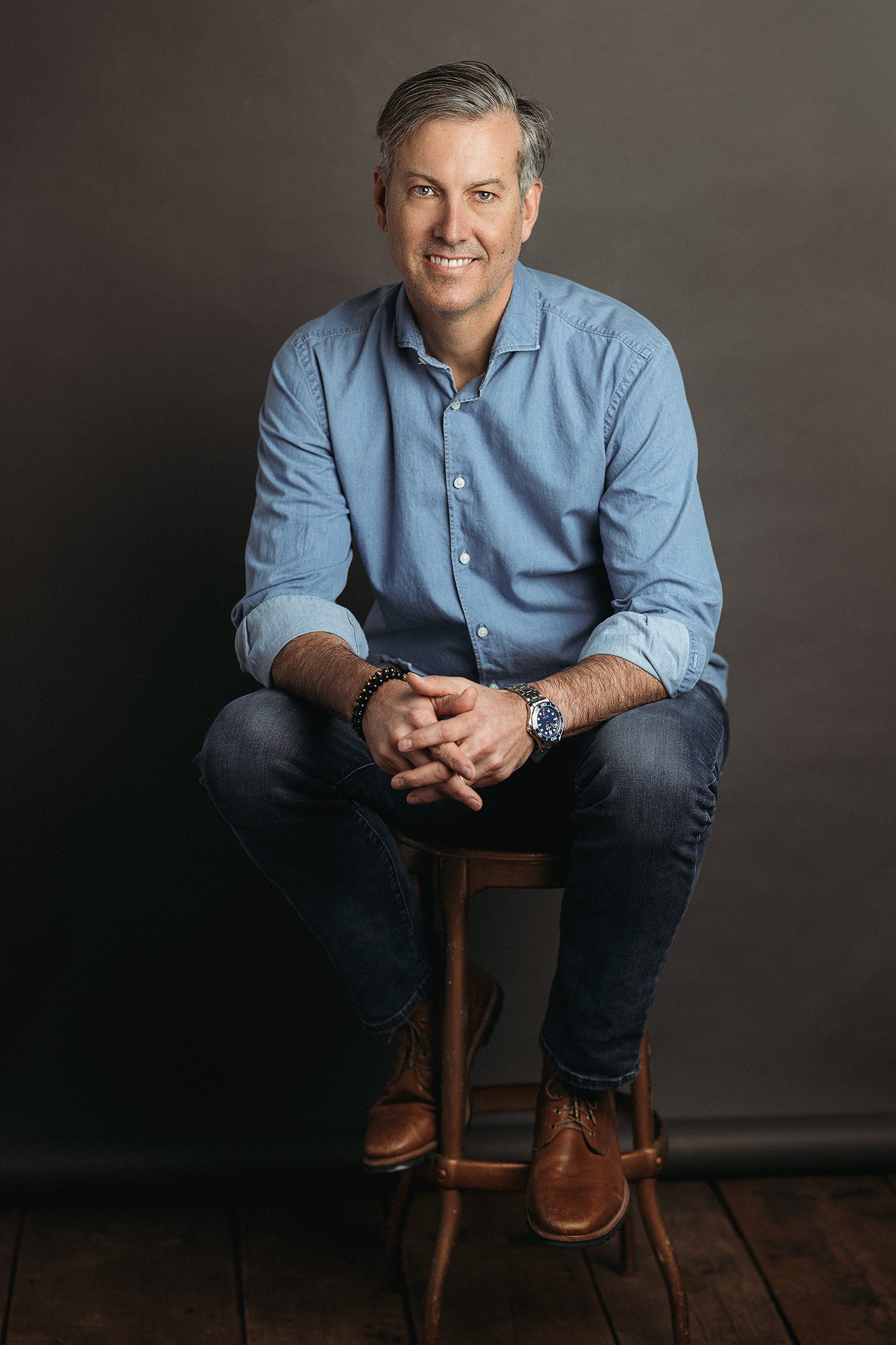 man wearing a blue denim shirt in jeans poses for headshot on grey backdrop