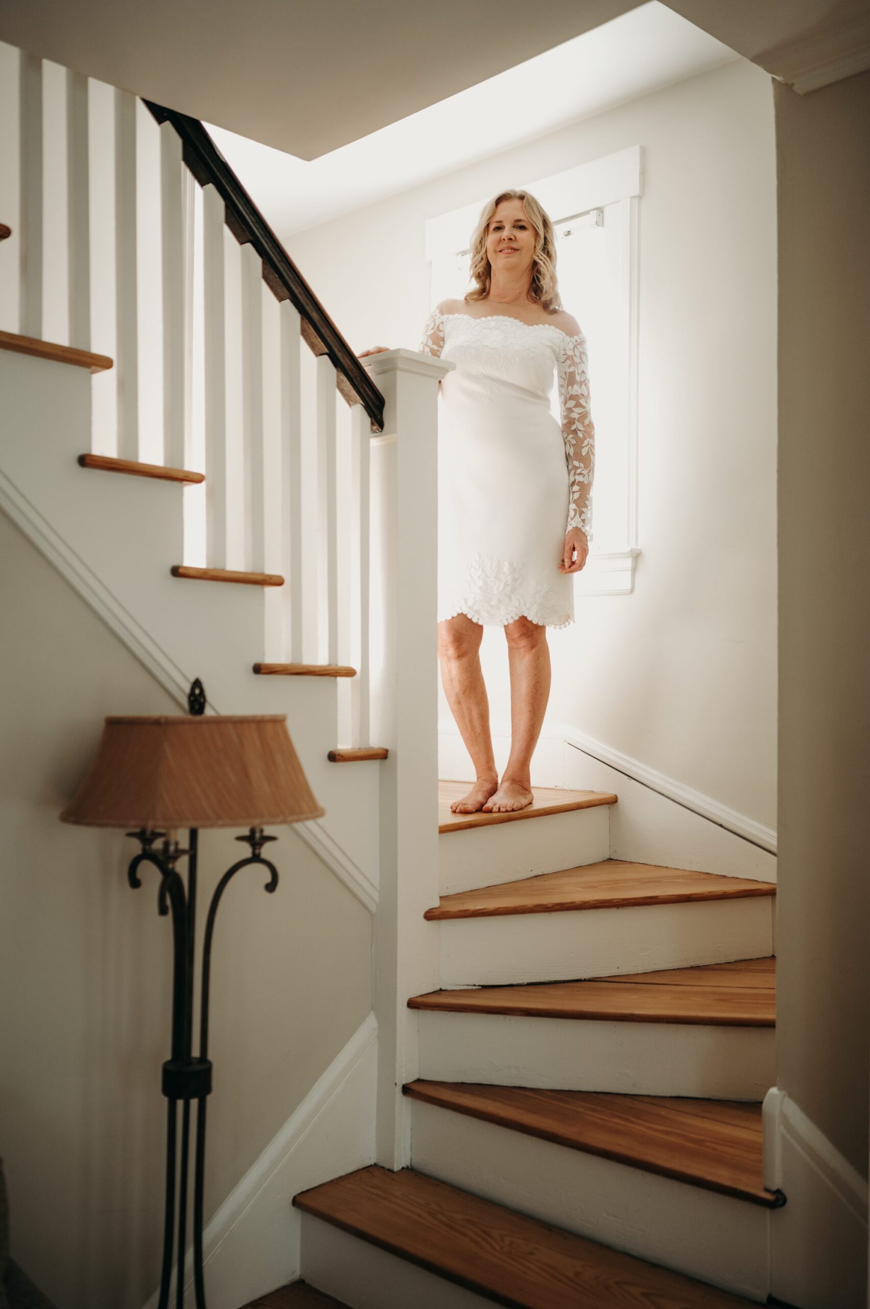 bride poses barefoot on the landing of her staircase with the light shining behind her